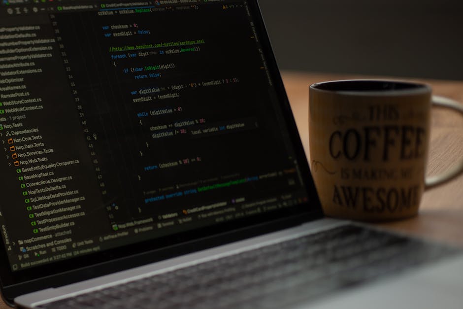 A close-up of a laptop computer displaying lines of colorful code on its screen, positioned on a wooden desk. To the right of the laptop, there is a ceramic mug with a beige background and black text