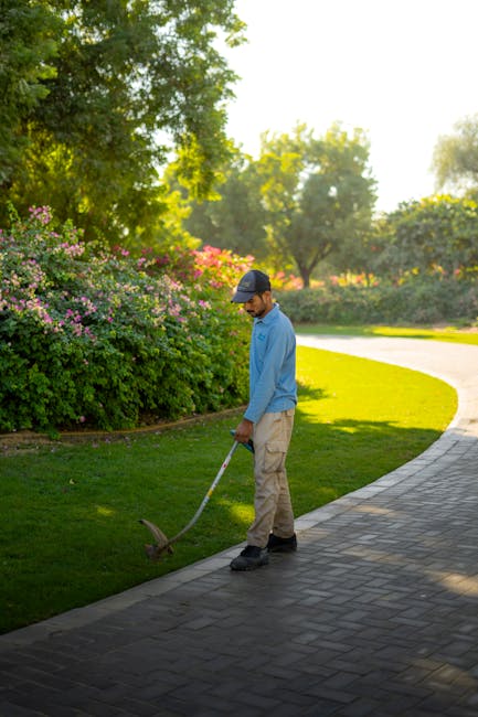 A gardener dressed in a blue shirt, beige trousers, and a cap is performing garden maintenance along a curved paved pathway. He is using a gardening tool to trim the edge of a well-manicured lawn, whi